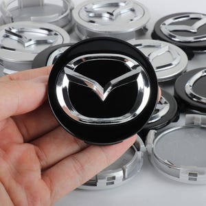 May include: A circular black wheel centre cap featuring a raised, polished chrome Mazda logo in the centre. The cap has a smooth, glossy finish on the front and a series of plastic clips on the underside for secure attachment to a vehicle wheel.