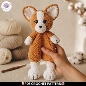 Puede incluir: Un peluche tejido a mano en forma de perro corgi, hecho con hilo suave de color marrón dorado y blanco. Presenta orejas puntiagudas, ojos de seguridad negros y una pequeña nariz negra. El peluche tiene una textura suave con patas blancas.