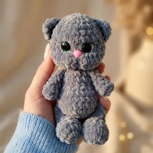 Puede incluir: Un pequeño gato de peluche tejido a mano con hilo de terciopelo suave de color gris oscuro. La figura presenta grandes ojos redondos de color negro brillante con reflejos verdes y una pequeña nariz de plástico lisa de color rosa claro.