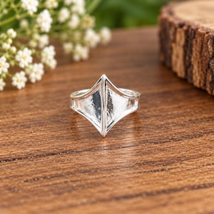 May include: A silver-toned ring featuring a prominent, pointed shield-like centerpiece with a smooth, reflective surface. The band is integrated into the geometric design, which tapers to a sharp vertical ridge at the center of the front face.