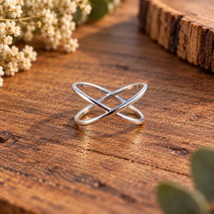 Può includere: Un anello in argento lucido caratterizzato da un design incrociato con due fasce che si intersecano formando una X. La superficie metallica è liscia e riflettente, catturando la luce per evidenziare le linee pulite della struttura dell'anello.