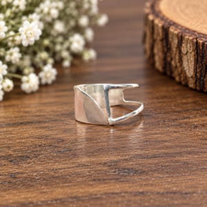 Op de afbeelding: Een zilveren ring met een brede, gladde en gepolijste band met een opvallend opengewerkt ontwerp. Het voorste gedeelte van de ring is massief en gebogen, terwijl het achterste gedeelte bestaat uit een dun, open metalen frame dat verbonden is.