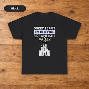 以下が含まれることがあります： 黒の半袖Tシャツ。白い文字で「SORRY, I CAN'T」、青いピクセル調の文字で「I'M PLAYING」、白い文字で「DREAMLIGHT VALLEY」と書かれており、その下に白い城のシルエットのグラフィックイラストが描かれています。