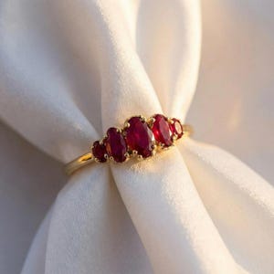 Puede incluir: Un anillo de oro con cinco rubíes rojos de talla ovalada dispuestos en fila. El rubí central es el más grande, con dos rubíes más pequeños a cada lado, montados en una banda de oro pulido que refleja la luz con un brillo cálido.
