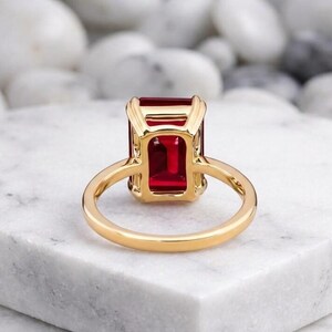 May include: A gold ring featuring a large, rectangular, emerald-cut red gemstone set in a four-prong basket setting. The band is smooth and polished, showcasing a vibrant, deep red stone that is held securely by the gold metal frame.