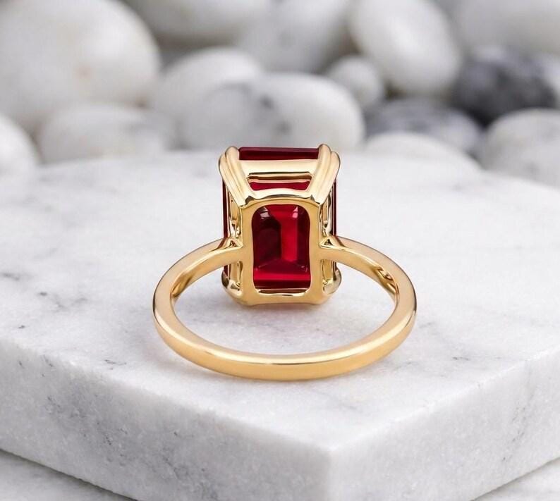 May include: A gold ring featuring a large, rectangular, emerald-cut red gemstone set in a four-prong basket setting. The band is smooth and polished, showcasing a vibrant, deep red stone that is held securely by the gold metal frame.