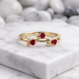 Könnte beinhalten: Ein goldener Ring mit einem Band aus zarten Metallblättern und drei runden, leuchtend roten Rubinen, die in Abständen auf der Oberseite gefasst sind. Der Ring hat eine polierte Oberfläche und eine geschwungene, organische Form.