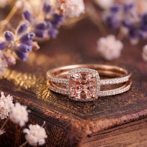 Könnte beinhalten: Ein Ring aus Roségold mit einem pfirsichfarbenen, kissenförmigen Mittelstein, der von einem Kranz aus kleinen weißen Diamanten umgeben ist. Die Ringschiene ist in zwei feine, mit Diamanten besetzte Reihen geteilt, die seitlich zusammenlaufen.