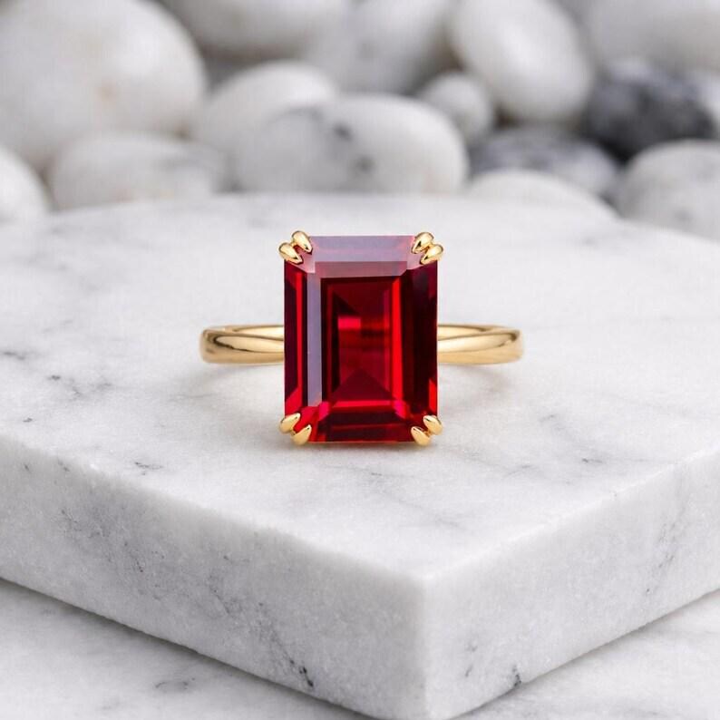 May include: A gold ring featuring a large, rectangular, emerald-cut vibrant red gemstone held in place by four prongs. The band is smooth and polished, showcasing a classic design that highlights the deep red color and clarity of the center stone.