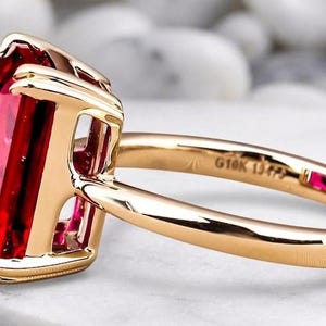 May include: A gold ring featuring a large, rectangular, faceted red gemstone held in a four-prong setting. The band is smooth and polished gold, with the text G10K 13475 engraved on the inner surface, accompanied by a small, square red accent stone.