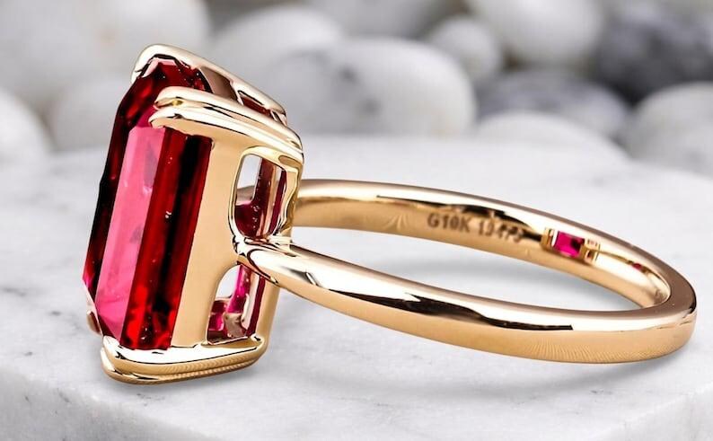 May include: A gold ring featuring a large, rectangular, faceted red gemstone held in a four-prong setting. The band is smooth and polished gold, with the text G10K 13475 engraved on the inner surface, accompanied by a small, square red accent stone.