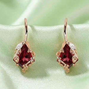 Könnte beinhalten: Ein Paar Ohrhänger aus Roségold mit einem drachenförmigen, tiefroten Edelstein in der Mitte. Jeder Ohrring ist mit kleinen, klaren, marquise-geschliffenen Steinen und zarten Metallblattmotiven um den zentralen roten Stein verziert.