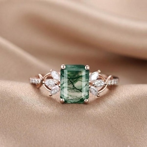 Könnte beinhalten: Ein Ring aus Roségold mit einem rechteckigen, im Smaragdschliff geschliffenen Moosachat-Mittelstein mit dunkelgrünen Einschlüssen. Die Ringschiene ist beidseitig mit drei weißen Steinen im Marquiseschliff besetzt, was ein symmetrisches Design ergibt.