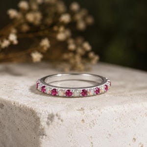 Könnte beinhalten: Ein silberner Ring mit einem abwechselnden Muster aus runden, facettierten rosa Edelsteinen und runden, milchig weißen Opalen, die in einer durchgehenden Reihe um den Umfang des Metallbandes gefasst sind und ein lebendiges, symmetrisches Schmuckstück bilden.