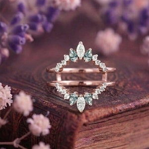 Peut inclure: Une bague double en or rose composée de deux anneaux incurvés reliés par un pont. Le design présente des pierres claires taille marquise au centre, flanquées de pierres vert mousse taille poire et de petites pierres rondes le long des anneaux.