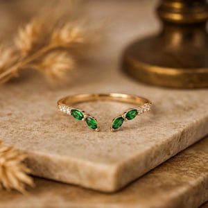 May include: A gold open-style ring featuring two marquise-cut green gemstones on each end, accented by small clear round stones along the band. The ring has a polished metallic finish and a delicate, symmetrical design with an open center gap.