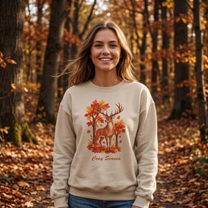 Peut inclure: Un sweat-shirt beige à col rond orné d'une illustration représentant un cerf au milieu d'arbres aux feuilles automnales orange et rouges. Le texte "Cosy Season" est imprimé en lettres cursives marron sous le motif sur le devant du vêtement.