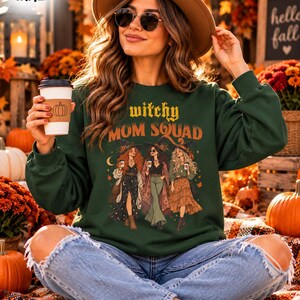 May include: A dark green long-sleeve sweatshirt featuring the text "witchy MOM SQUAD" above a graphic illustration of three figures wearing hats and bohemian clothing. The sweatshirt is paired with light blue distressed denim jeans.