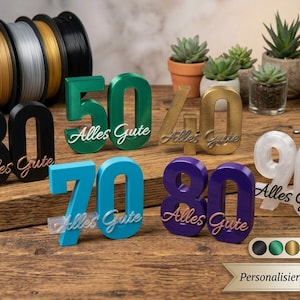 Op de afbeelding: Een verzameling 3D-geprinte cijferborden in diverse kleuren, waaronder zwart, groen, goud, blauw, paars en wit. Elk bord toont het cijfer en de cursieve tekst Alles Gute. De cijfers 30, 40, 50, 70, 80 en 90 zijn weergegeven.