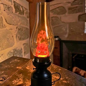 May include: A black metal oil lamp base with a clear glass chimney containing a glowing orange and red flame-shaped figure with two white eyes and a small mouth, resembling a character from a fantasy animation, set on a dark surface.