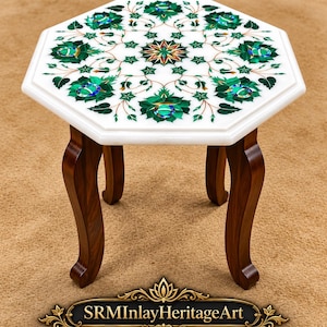 Könnte beinhalten: Ein achteckiger Tisch aus weißem Marmor mit kunstvollen Einlegearbeiten aus grünen und bunten Steinen in Blumenmustern auf der Oberfläche, getragen von vier dunkelbraunen Holzbeinen. Der Text SRMInlayHeritageArt ist unten sichtbar.