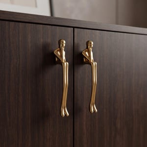 Può includere: Due maniglie per armadietto in metallo color oro a forma di figure stilizzate sedute con le gambe a penzoloni. Ogni maniglia presenta una finitura metallica liscia ed è montata verticalmente su una porta di armadietto in legno marrone scuro.