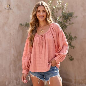 Puede incluir: Una blusa de color melocotón de manga larga con un patrón texturizado de cuadrícula, cuello redondo y cierre de lazo frontal. La prenda presenta mangas sueltas y abullonadas con puños fruncidos, combinada con pantalones cortos de mezclilla azul con dobladillo deshilachado.