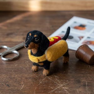May include: A needle-felted black and tan dachshund figurine wearing a yellow hot dog bun costume with a red stripe representing ketchup. The small dog has dark eyes, a black nose, and stands on four paws with a raised tail.