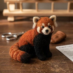 May include: A needle-felted red panda figurine with a rust-orange back, a long striped tail, and black fur on the chest and legs. The creature has white facial markings, pointed ears, and dark eyes, sitting in a seated position on a wooden surface.
