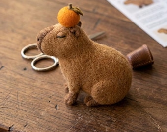 Op de afbeelding: Een bruin naaldgevilte capybara-figuurtje met gesloten ogen, met een kleine oranje vrucht met een groen blaadje op het hoofd. Het figuurtje staat bij een zilveren metalen schaar en een bruine vingerhoed op een houten ondergrond.