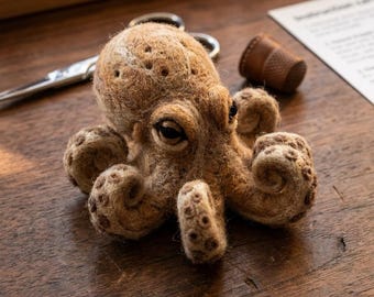 Op de afbeelding: Een naaldgevilte octopusfiguur gemaakt van beige wolvezels. Het wezen heeft een afgeronde kop met kleine inkepingen, donkere glanzende ogen en acht gekrulde tentakels met zichtbare cirkelvormige zuignapdetails aan de onderzijde.
