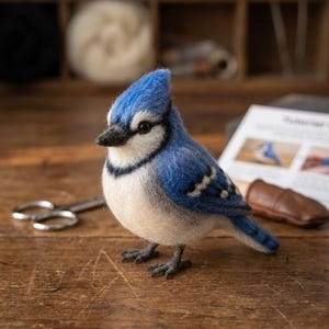 Peut inclure: Une figurine de geai bleu en laine feutrée à l'aiguille avec une crête bleue vive, une poitrine blanche et des marques faciales noires. L'oiseau possède un bec sombre et de petites pattes foncées, avec une finition en laine douce et texturée.