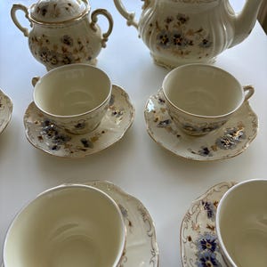 Könnte beinhalten: Ein cremefarbenes Porzellan-Teeservice bestehend aus einer Teekanne mit Deckel, einer Zuckerdose mit Deckel und vier Teetassen mit passenden Untertassen. Jedes Teil ist mit einem zarten Blumenmuster in Blau- und Goldtönen sowie Goldrand verziert.