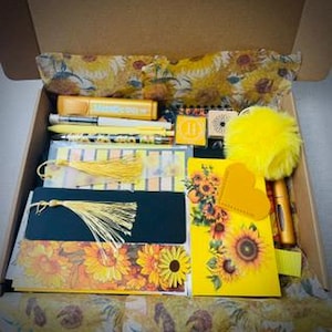 May include: A stationery gift box filled with sunflower-themed items including a yellow highlighter, pens, bookmarks with yellow tassels, a notebook with a sunflower cover, a yellow heart-shaped tag, and a bright yellow fluffy pom-pom accessory.