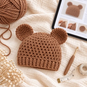 Puede incluir: Un gorro de ganchillo marrón con dos orejas de oso redondeadas en la parte superior. El gorro está confeccionado con un patrón de punto texturizado y un borde acanalado. Se muestra junto a un ovillo de lana marrón, un ganchillo y una cinta métrica.