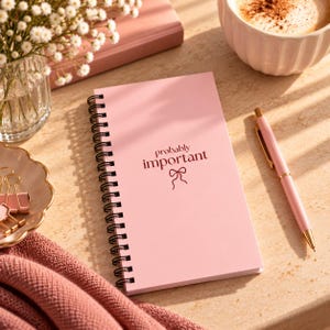 May include: A light pink spiral-bound notebook with black wire binding. The cover features the dark red text "probably important" above a small line-art bow icon. The notebook is positioned next to a matching light pink pen with gold accents.