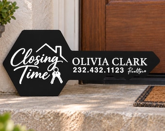 May include: A black, key-shaped sign featuring the white text "Closing Time" with a house icon and two keys. To the right, the white text reads "OLIVIA CLARK", "232.432.1123", and "Realtor". The sign has a hexagonal head and a pointed blade end.