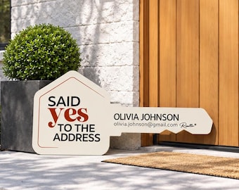 May include: A cream-colored, key-shaped sign featuring the text "SAID YES TO THE ADDRESS" in black and red lettering, along with the name "OLIVIA JOHNSON" and an email address printed on the blade of the key, displayed on a concrete porch.