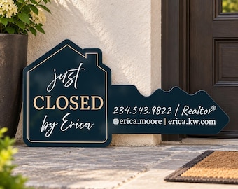 May include: A dark blue, house-shaped sign in the form of a key with gold text reading "just CLOSED by Erica", "234.543.9822 / Realtor", and social media handles. The sign features a thin gold border and is displayed on a stone porch.
