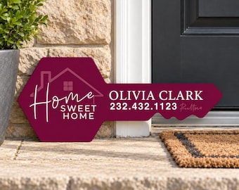 May include: A burgundy key-shaped sign featuring a white house outline and the text Home Sweet Home, Olivia Clark, and 232.432.1123. The sign is positioned on a stone porch next to a dark door and a green plant in a gray planter.