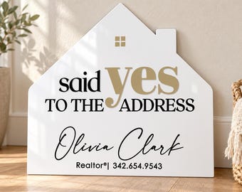 May include: A white house-shaped sign featuring the gold and black text "said yes TO THE ADDRESS" with the name "Olivia Clark" and "Realtor® | 342.654.9543" printed below. The sign includes a small gold square window icon at the top.