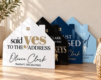 May include: A row of four house-shaped signs in white, black, navy, and light blue. The white sign features the text 'said yes TO THE ADDRESS' and 'Olivia Clark Realtor 342.654.9543'. Other signs display realtor names, keys, and contact details.