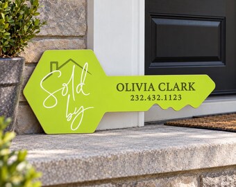 May include: A lime green, key-shaped sign featuring a white house icon and the script text Sold by, along with the printed name OLIVIA CLARK and the phone number 232.432.1123 in dark gray lettering, displayed on a stone porch step.