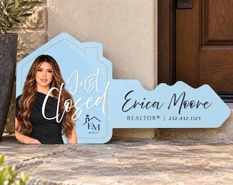 May include: A light blue, house-shaped sign featuring a photograph of a person with long brown hair wearing a black top. The text reads Just Closed, EM Realty, and Erica Moore REALTOR with the phone number 232.432.1123 printed in black lettering.