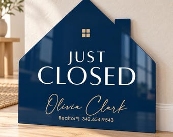 May include: A house-shaped sign in dark blue with a glossy finish. The text reads JUST CLOSED in white, with the name Olivia Clark and the text Realtor 342.654.9543 in gold. A small gold square window icon is centered near the roof peak.