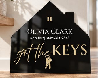 May include: A house-shaped black sign with gold text reading Olivia Clark, Realtor, 342.654.9543, and got the KEYS, featuring a small gold window icon and a gold key graphic. The sign has a glossy finish and stands on a wooden surface.