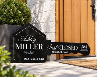May include: A black, key-shaped sign featuring white text that reads Ashley MILLER Realtor, Just CLOSED, ashM.real, and 234.211.5432, along with an AM REALTY logo. The sign is positioned on a concrete surface near a wooden door.