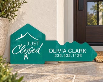 May include: A teal, key-shaped sign featuring a white house icon, the text "JUST Closed" with a small key graphic, and the name "OLIVIA CLARK" followed by the phone number "232.432.1123" printed in white lettering on the surface.