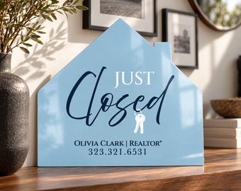 May include: A light blue house-shaped sign with the text "JUST Closed" in dark blue and white, featuring a white key icon. Below, the text reads "OLIVIA CLARK | REALTOR" and the phone number "323.321.6531" in dark blue lettering.