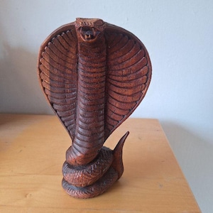 Op de afbeelding: Een donkerbruin houten sculptuur van een cobra in een rechtopstaande, defensieve houding met een gespreide kap. Het houtsnijwerk is voorzien van gedetailleerde horizontale ribbels langs de nek en het lichaam, met een open bek die kleine slagtanden toont.